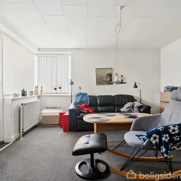 En stue med en sort lædersofa og sofabord i træ. En grå lænestol står ved siden af. Mange flyttekasser er stablet i rummet, som har hvide vægge og et tæppebelagt gulv.