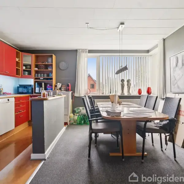 Et spisebord med dækkede tallerkener står i en spisestue med røde køkkenskabe til venstre. Et stort vindue giver udskig til omgivelserne, omgivet af malerier og indretning.