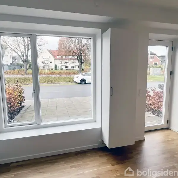 Et rum med trægulv og store vinduer, der viser en gade med parkerede biler og hække. Udenfor ses huse og træer.
