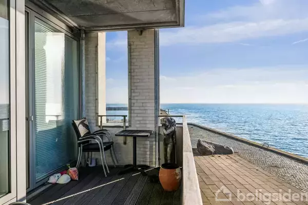 En altan med to stole og et bord står mod havet. Murstensvægge omgiver området, mens en klar, blå himmel udgør baggrunden.