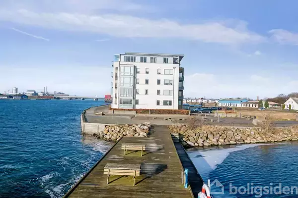 En hvid bygning står ved kysten, omgivet af vand og sten, med en træbro i forgrunden. To bænke står på broen, og himlen er klar.