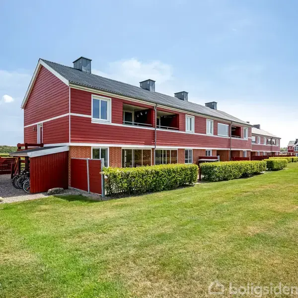 Rødt rækkehus med flere etager, omgivet af græsplæne og bede med buske. En bil og cykler står parkeret på grusvej ved siden af huset. Klar himmel i baggrunden.
