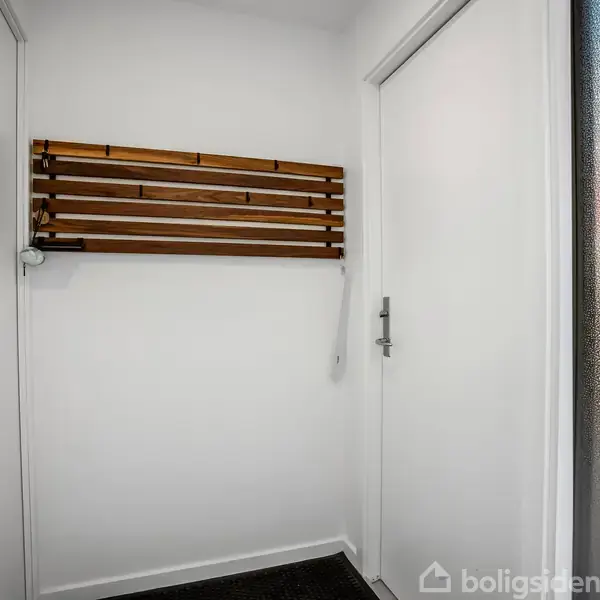 En vægmonteret træhylde hænger stille på en hvid væg. Rummet har glatte vægge og døre, med en delvist mat glasdør til højre, hvilket antyder en entré eller gang.