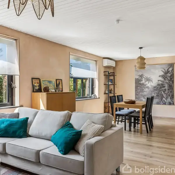 En lys stue med en beige sofa foran to vinduer. Sofaen har farverige puder, og der er et spisebord med fire stole i baggrunden. En vægdekoration forestiller palmetræer.