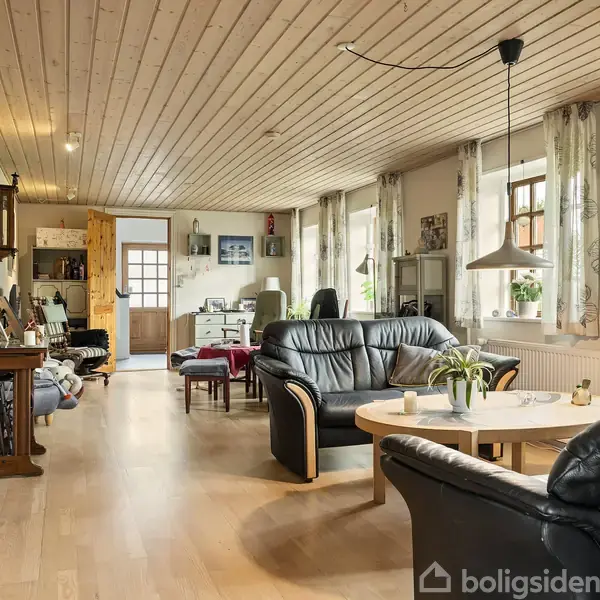 En stue med sorte lædersofaer og et rundt sofabord prydet med planter. Rummet har trægulv og træloft, vinduer med gardiner, og lys hænger fra loftet.