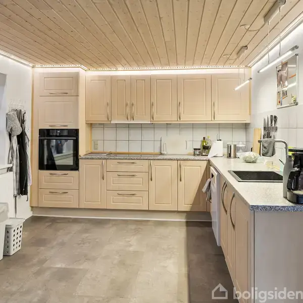 En rummelig, moderne køkken med lyse træskabe og integreret ovn. Kaffemaskine står på bordet ved siden af vasken. Et køleskab er til venstre, omgivet af hængende jakker.