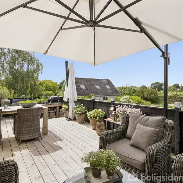 En terrasse med flettede stole og bord under en stor parasol. Omgivet af planter og træer, med udsigt over en grøn have og et tag på et hus i nærheden.