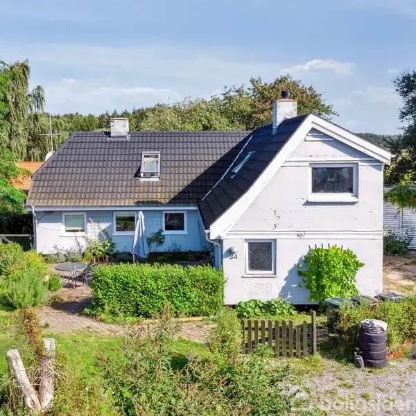 Hus med sort tag står i frodig have, omgivet af buske og træer. En lys og solrig dag kaster skygger på facaden. En sti fører til hoveddøren.