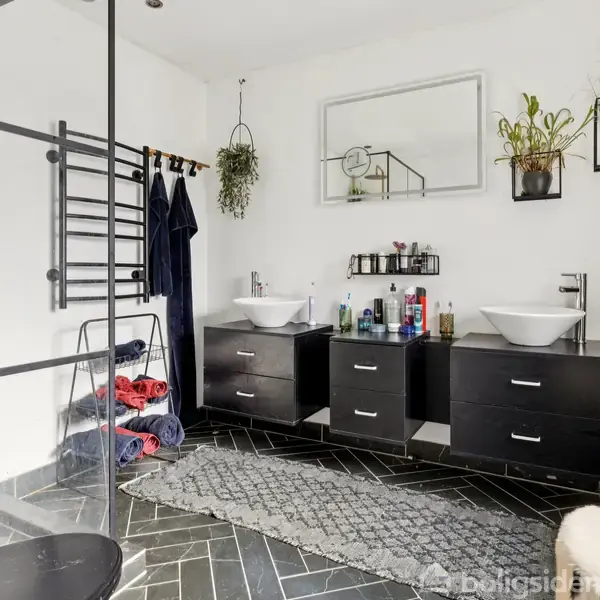 Toilet med sorte skabe under to håndvaske, planter på væggen, håndklæder på en stige, og et tæppe på sort gulv. Store spejle hænger over vaskene.