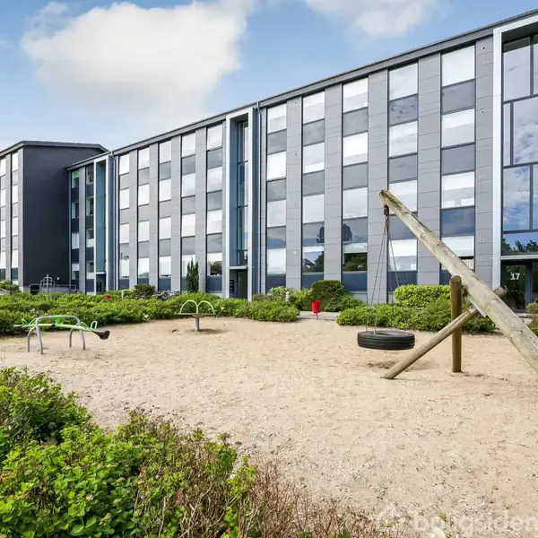 Legeplads med en vippe står i et sandet område foran en moderne, fleretagers bygning med glasfacade og mørke paneler. Grønne buske omkranser legepladsen.