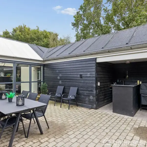 Udendørs terrasse med sort bord og seks stole. To liggestole står ved den mørke træfacade. Et udekøkken er placeret under halvtag. Omgivet af træer og klart vejr.