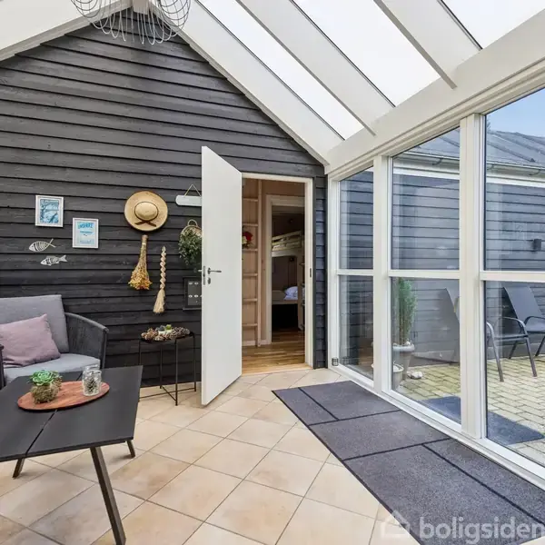 Sort træhus med udendørs terrasse, hvor et bord og stole står på lyse fliser. Store vinduer viser ind i huset. Omgivet af grønne træer under en blå himmel.