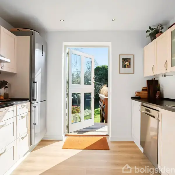 Køkken med lyse skabe og sort bordplade fører ud til åben dør, som giver udsigt til grøn have i sollys. Vægge har enkelte dekorationer.