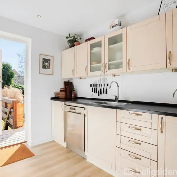 En lys køkkenvask med hvide skabe og sorte greb, omgivet af planter, står i et rum med åbne franske døre, der leder ud til en solrig terrasse med udsigt over en have.