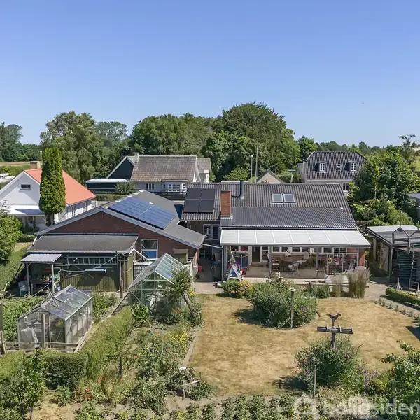 Hus med solpaneler på taget ligger i villakvarter. Haven rummer drivhuse, græsplæne og forskellige planter. Omgivet af andre boliger og træer, på en solrig dag.