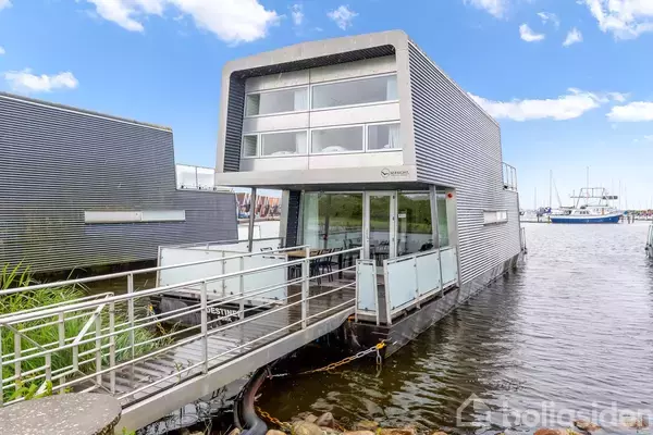 Husbåd flyder stille ved kajen i en havn, omgivet af vand og andre både. Bygningen har moderne arkitektur med store vinduer og en bro, der forbinder til land.