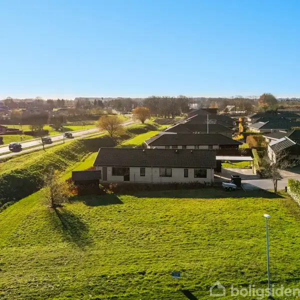 Et hus står stille i en forstad med grønne græsplæner omkring. Baggrunden viser en vej med biler og træer under en klar blå himmel.