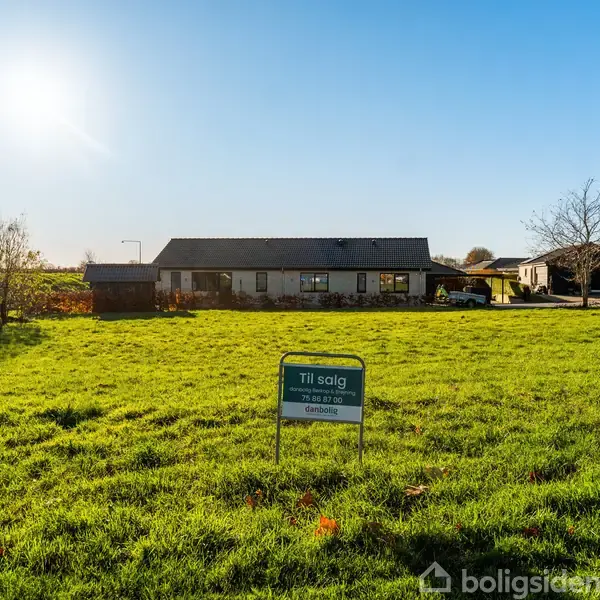 Et hus ligger stille på en stor, grøn græsplæne under en klar blå himmel med solen skinnende. Et skilt foran grunden viser "Til salg" med et telefonnummer.