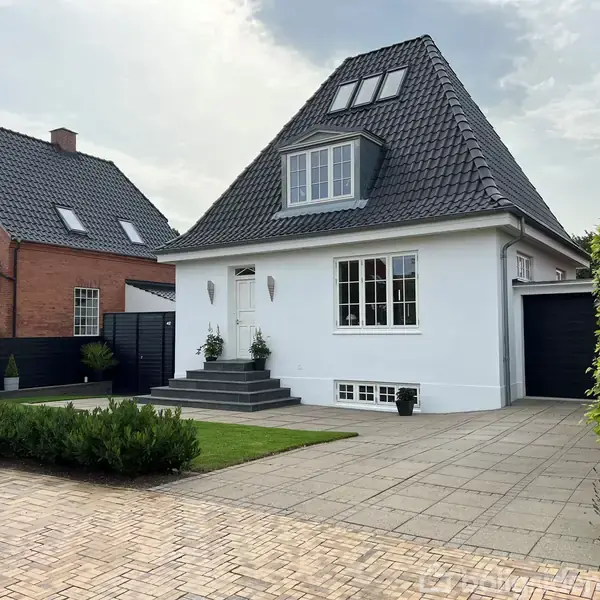 En hvid, firkantet villa med sort tag står på en flisebelagt indkørsel. Villaen har en sort dør, trapper op til indgangen, og vinduer med sprosser. En rød murstensbygning ses til venstre.