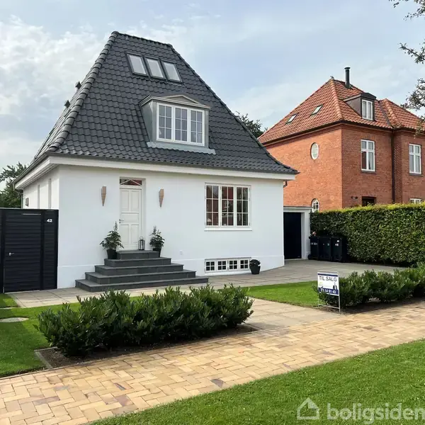 Hvidt hus med mørkt tag og kvist, omkranset af grøn hæk. Det står ved siden af et rødt murstenshus. Indgang med trappe. Skilt foran huset med teksten "TIL SALG home".