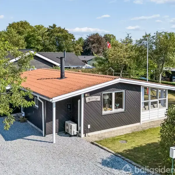 Et sommerhus med rødt tag står i en have med græsplæne og stenbelagt indkørsel. En flagstang ses i baggrunden. Skilt på huset: "12 Mortenstrup."