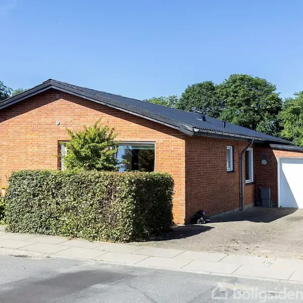 Rødt murstenshus med sort tag ligger ved siden af en flisebelagt indkørsel og garage. Hækken omgiver forhaver. Træer ses i baggrunden under en klar blå himmel.
