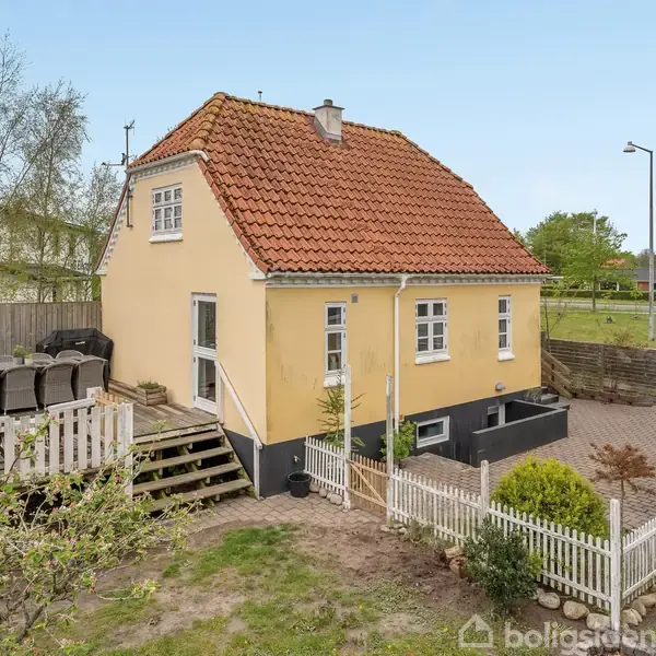 Et gulmalet hus med rødt tegltag og hvide vinduer står i en have med et hvidt stakit. Der er terrasse med møbler. Omgivelserne har grønne træer og en vej.