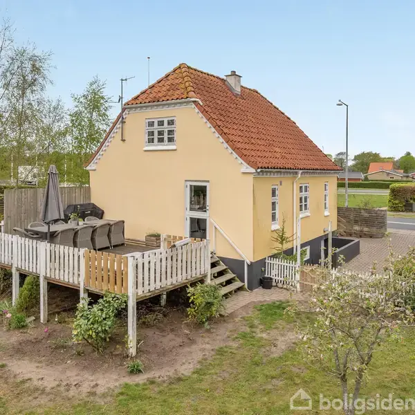 Et gult hus med rødt tag står ved en hævet træterrasse, der har havemøbler og en parasol. Omkring er der græsplæne og træer i et parcelhuskvarter.