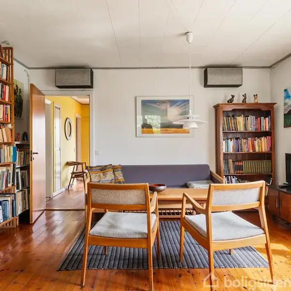 To lænestole vender mod en sofa i en stue med parketgulv. Bogreoler fylder rummets vægge, mens et tæppe ligger under møblerne. Vinduerne er højreorienterede og giver naturligt lys.