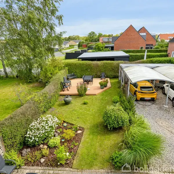 En velplejet have med blomsterbede og hæk omkranser en terrasse med spisebord og stole. Til højre er en grusbelagt indkørsel med to biler parkeret under et halvtag i et villakvarter.