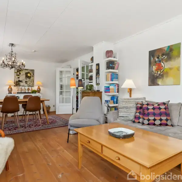 En stue med grå sofa og træbord foran, dekoreret med farverige puder. Ved siden af står en bogreol og lampe. I baggrunden ses et spisebord med seks stole under en lysekrone. Malerier hænger på væggen.