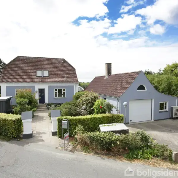 Gråmalet hus med rødt tegltag, omgivet af hække og buske, står ved siden af en garage. Foran er en stenbelagt indkørsel med parkeret bil. Skyede himmel i baggrunden.