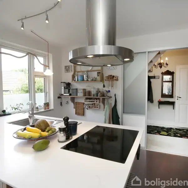 Et moderne køkken med en hvid køkkenø fungerer som arbejdsområde. Frugt er placeret i en skål, og en emhætte hænger over kogepladerne. I baggrunden ses døre og en gang.