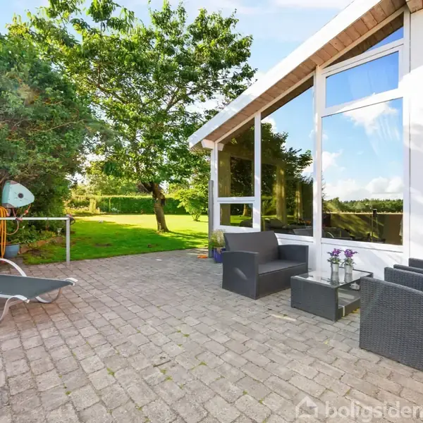 Terrassemøbler står på en flisebelagt terrasse foran et moderne hus med store vinduer. En liggestol og to stole med et bord står i solen, omgivet af grønne træer og græsplæne.