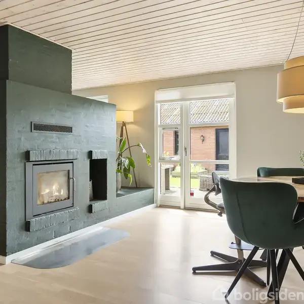 Brændeovn indbygget i sort murstensvæg fungerer som rumdeler, omgivet af moderne spisestue med grønne stole og et rundt bord. Store vinduer giver udsigt til haven.