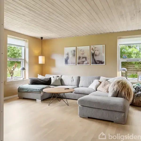 Stue med en stor, grå sofa og runde sofaborde. Væggene prydes af kunst, og der er store vinduer med udsigt til en have. Blød belysning og dekorative hylder.