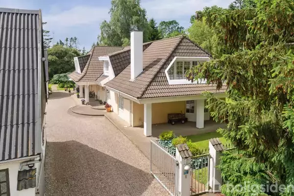 Et stort hus med gavl og kamin står stille midt i en grøn have. Grusvej fører til indgangsport, omkranset af træer og buske.