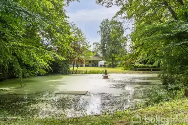En lille overdækket dam ligger fredfyldt omgivet af tætte træer. I baggrunden ses et hvidt hus, der tilføjer en idyllisk atmosfære til det grønne landskab.