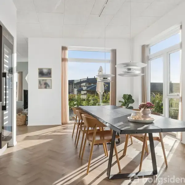 Spisebord med stole står i et lyst rum med store vinduer, der giver udsigt til en have. På bordet er en dekoration. Kunst hænger på væggen, træparketgulv skaber hygge.
