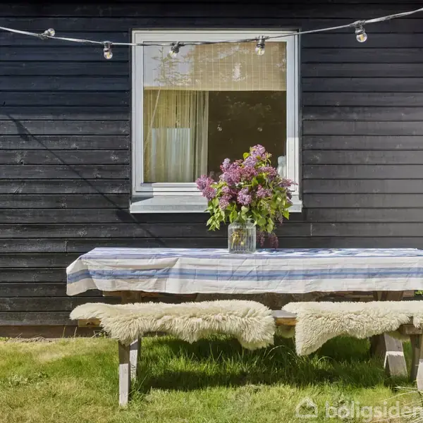 Langbord dækket med stribet dug står foran sort trævæg. Buket af lilla blomster midt på bordet. Lyskæde hænger over bordet. Græsplæne omgiver arrangementet.