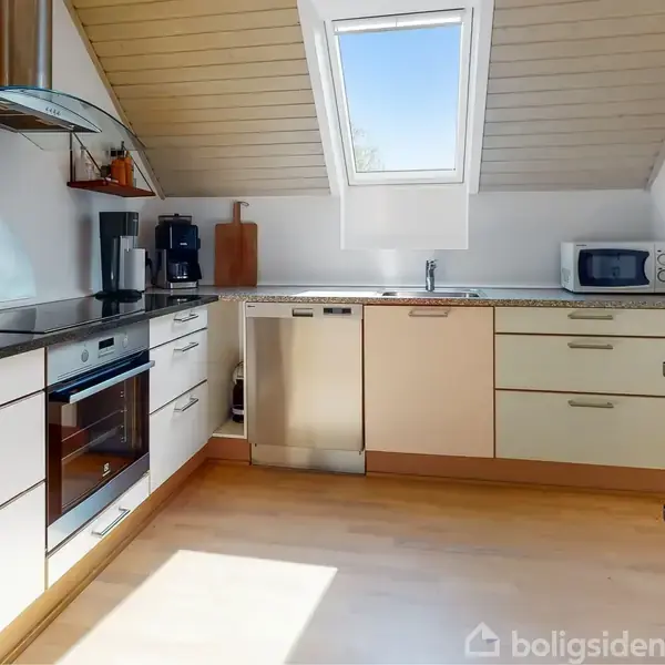 En moderne køkkenindretning med hvide skabe og stålknopper, udstyret med ovn, kogeplade, opvaskemaskine og mikroovn. Et stort ovenlysvindue giver naturligt lys. Flasker står på en køkkenvogn.