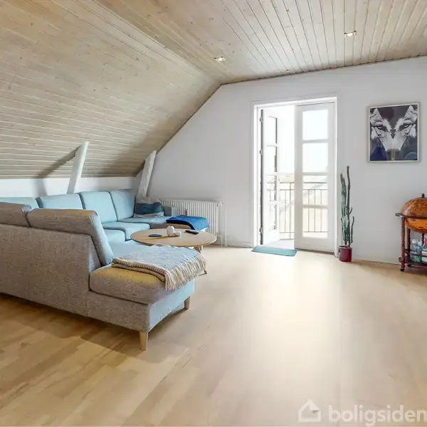 En grå sofa står i en stue med skråloft og trægulv. En dør fører til en balkon, fladskærms-TV hænger på væggen, og en globusbar står ved siden af en plante.