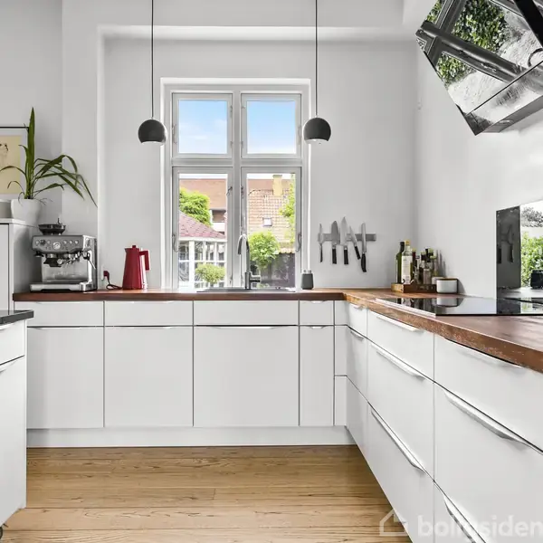 Et moderne køkken med hvide skabe, træbordplader, og et espresso-apparat ved siden af en rød elkedel. Vindue med udsigt til en have, omgivet af grønne træer.