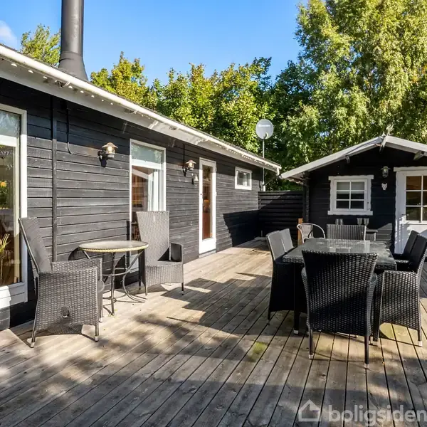 Sort sommerhus med træterrasse, hvor sorte kurvemøbler står opstillet omkring runde borde. Omkring er frodig beplantning og blå himmel, hvilket skaber en afslappet, udendørs atmosfære.