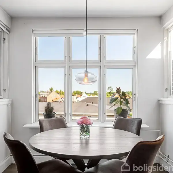 Rundt spisebord med fire stole står foran stort vindue. En vase med blomster pryder bordet. Lysekrone hænger over bordet. Billederammer og planter dekorerer rummet.