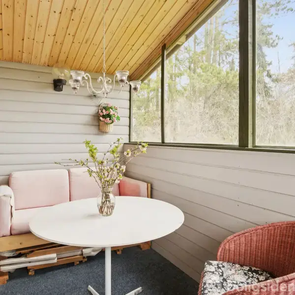 Sofa og bord med blomst i vase står i en lukket balkon med trævægge og loft. Rød lampe og hængeplante tilføjer hjemlig atmosfære, mens store vinduer viser skovudsigt.