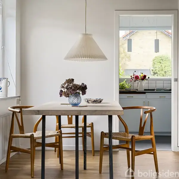 Et spisebord med fire træstole står i et lyst rum. En vase med blomster pryder bordet. I baggrunden ses et køkken med en åben dør.