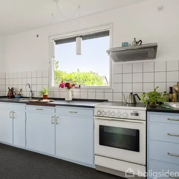 Lyst køkken med lyseblå skabe, sort bordplade, og hvidt komfur. En vask er til venstre med planter og frugter ovenpå. Fliser på væggen. Vindue med udsigt til grønne omgivelser.