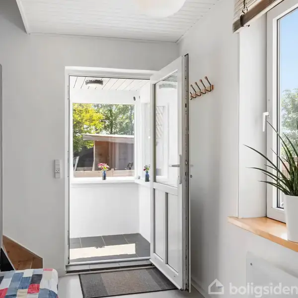 Indgangsområde med hvid dør åben mod en lys veranda. Til venstre ses en bænk med farverigt tæppe. Til højre står en plante i vindueskarmen. Udenfor er der grønt.