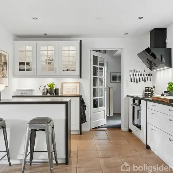 Et lyst køkken med hvidt interiør og sorte detaljer. To barstole står ved en køkkenø. På væggene hænger knive, og der er planter og en brødrister.